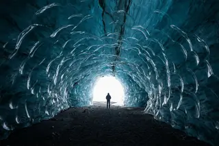 The inside of a glacier ice cave glowing deep blue with a small figure at the entrance.