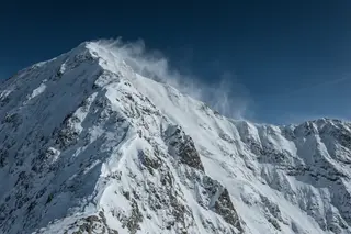 A knife-edge snow ridge with plumes of wind-blown snow against a deep blue sky.