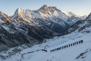 A line of tiny climbers crossing a vast snow slope beneath Himalayan peaks at dawn.