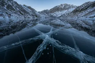 Clear black lake ice with white cracks reflecting snowy mountains.